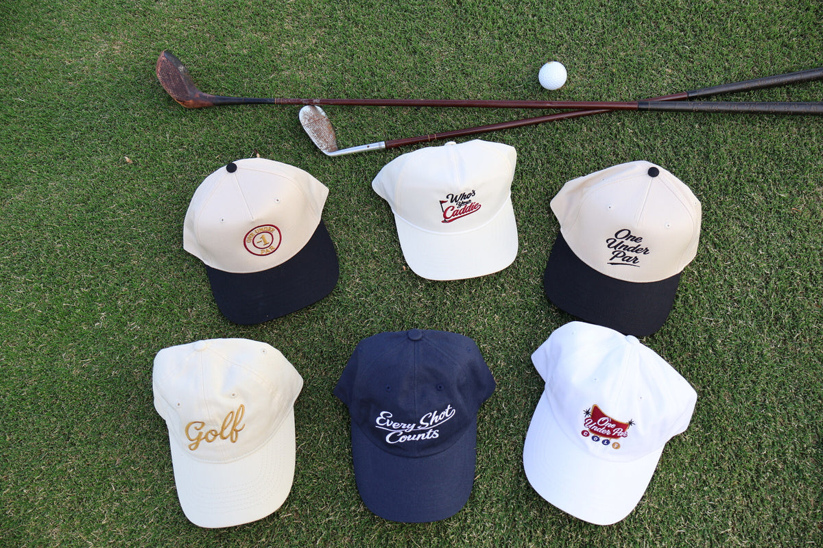 One Under Par golf hats arranged with vintage clubs and a golf ball on the course